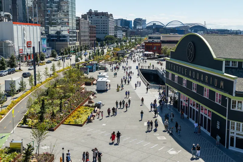 Seattle's Waterfront Park, as seen from atop the Overlook Walk looking south. The Seattle Aquarium's Pier 59 building is featured to the right, with pathways, bike paths, and green spaces along the park.